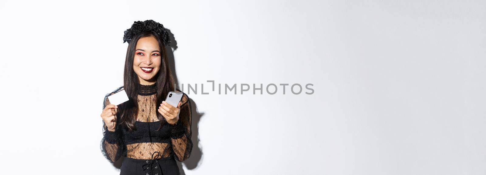 Image of beautiful asian woman in gothic lace dress and black wreath, looking aside pleased and smiling, holding mobile phone with credit card.