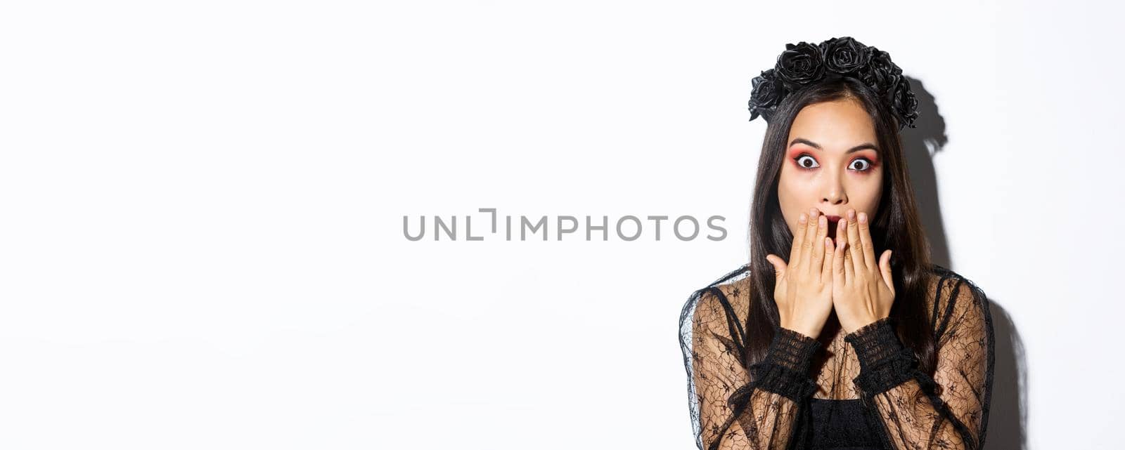 Close-up of shocked asian woman in gothic lace dress and wreath gasping, cover mouth and stare at camera amazed, standing over white background.