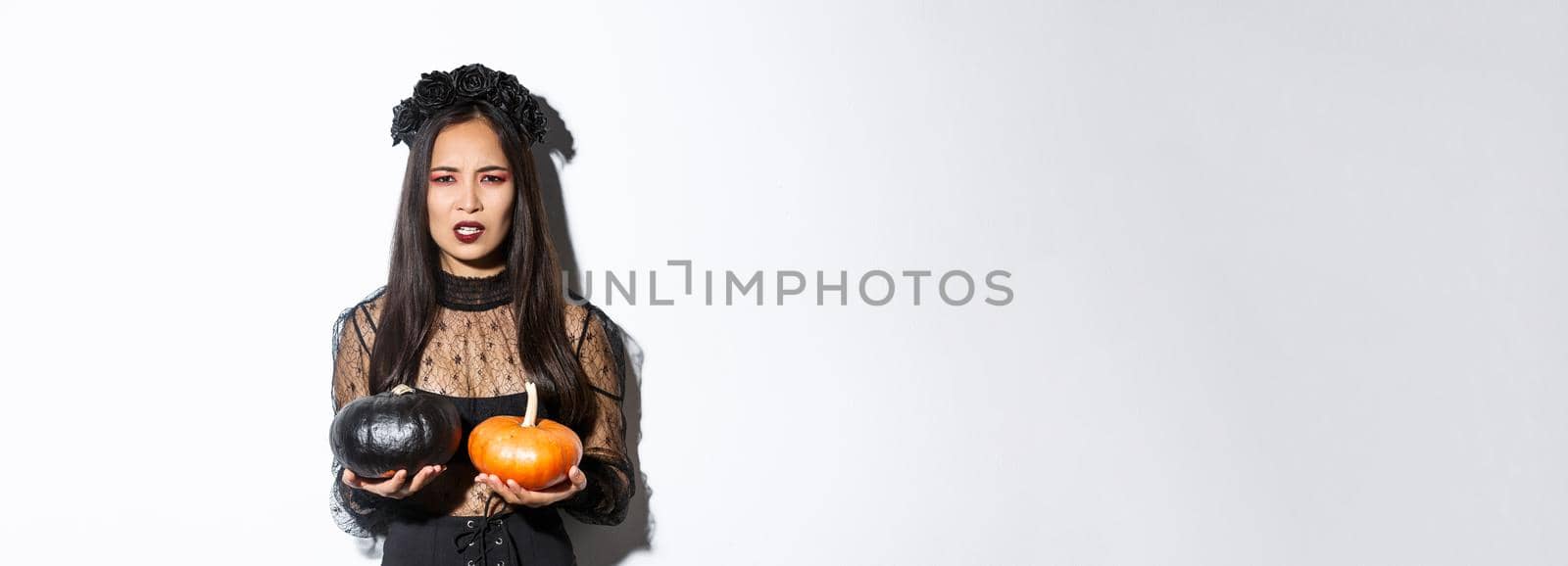 Image of grimacing angry witch throwing pumpkins, girl celebrating halloween, standing over white background by Benzoix