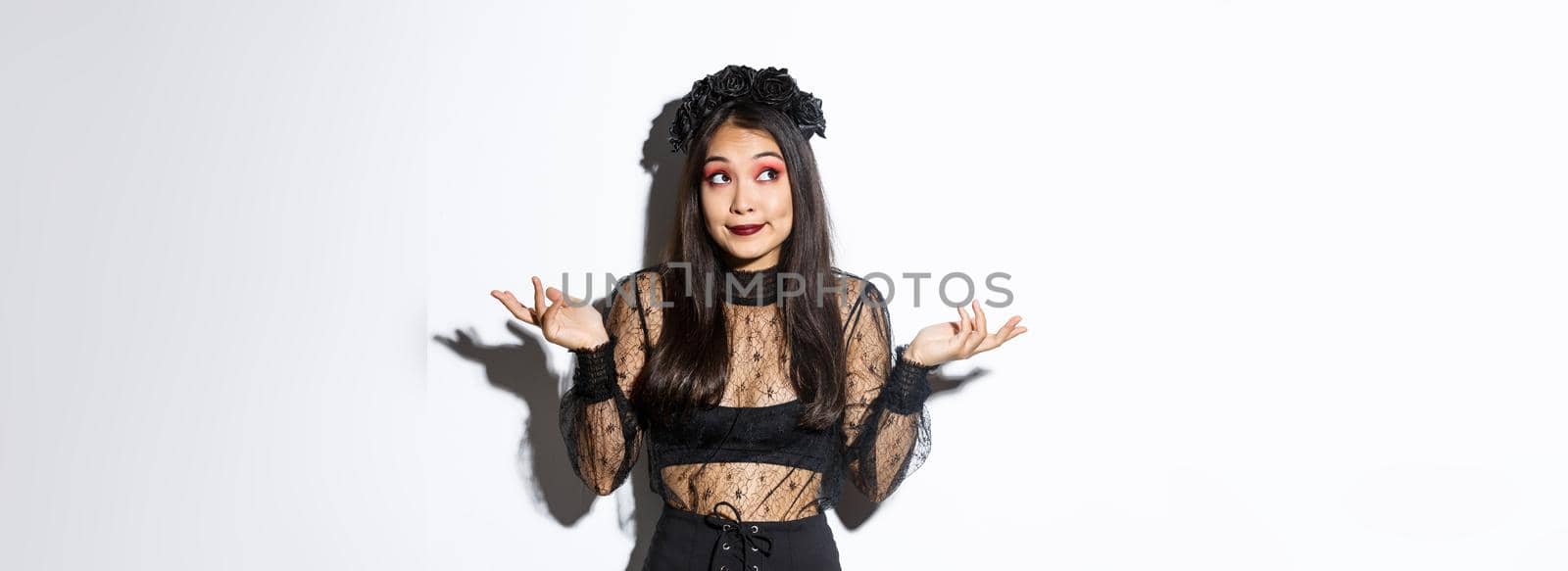Image of clueless asian girl dressed-up as a witch, shrugging and smiling, looking upper left corner at halloween banner or your logo, standing over white background in gothic dress.
