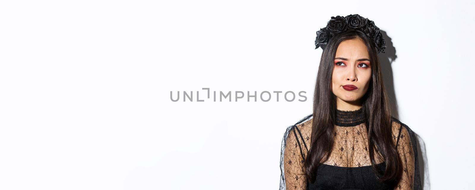 Close-up of perplexed asian woman in wicked witch costume, looking at upper left corner doubtful or indecisive, standing over white background.