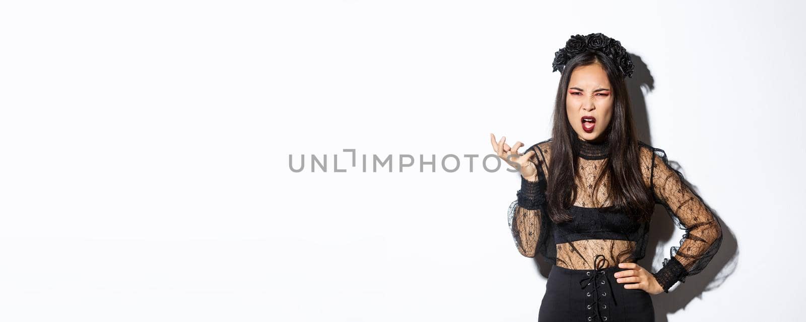 Frustrated and angry young asian woman in witch dress arguing with someone, looking pissed-off and bothered, shaking hand and looking scary at camera, standing over white background by Benzoix