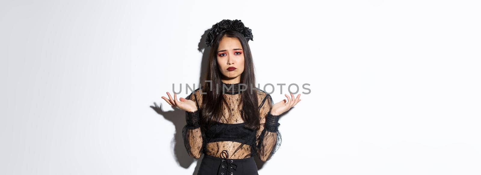 Clueless pensive asian girl in witch costume looking indecisive, shrugging with hands spread sideways, looking sad over white background.