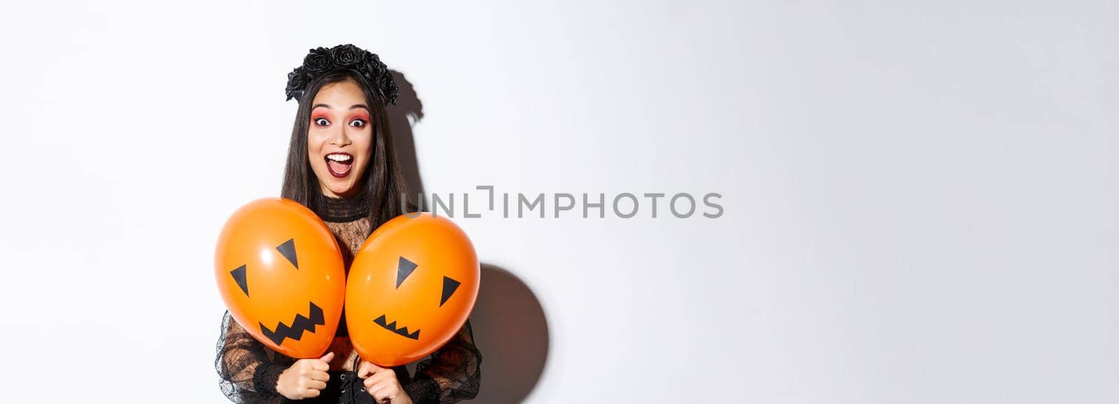 Image of asian girl in evil witch costume holding two orange balloons with scary faces, celebrating halloween, standing over white background by Benzoix