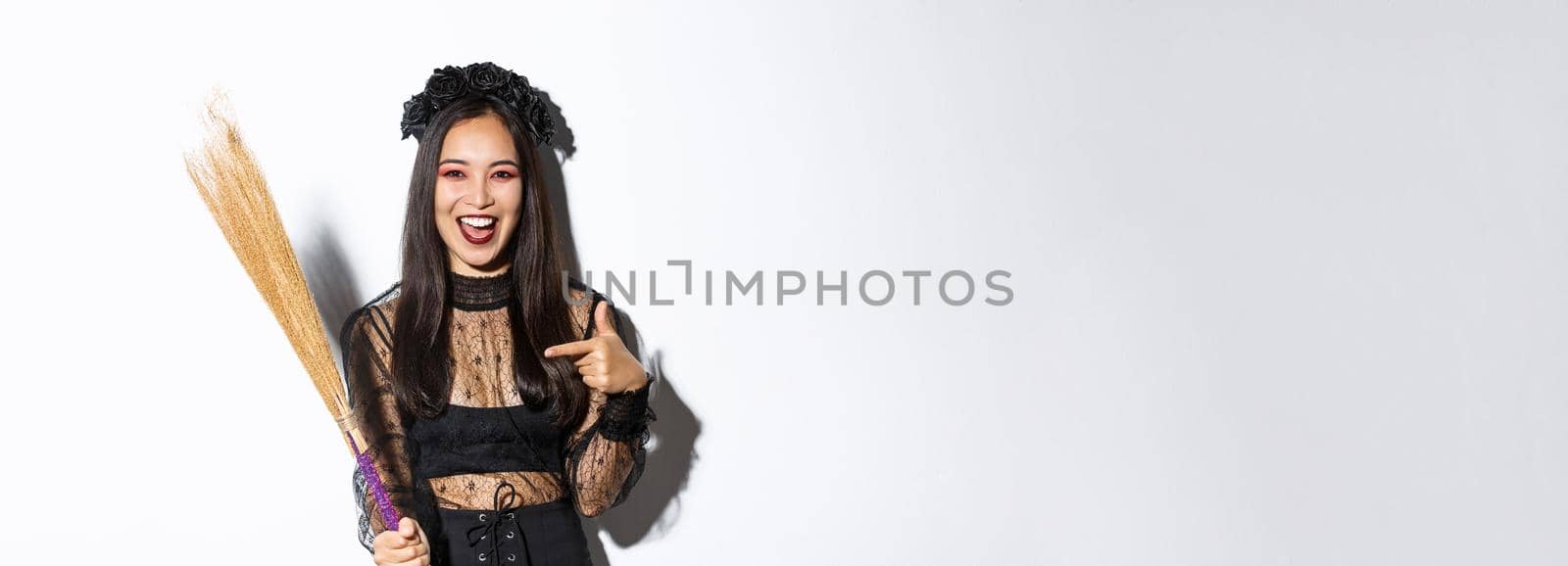 Portrait of attractive woman in witch costume celebrating halloween, pointing at broom, standing over white background by Benzoix