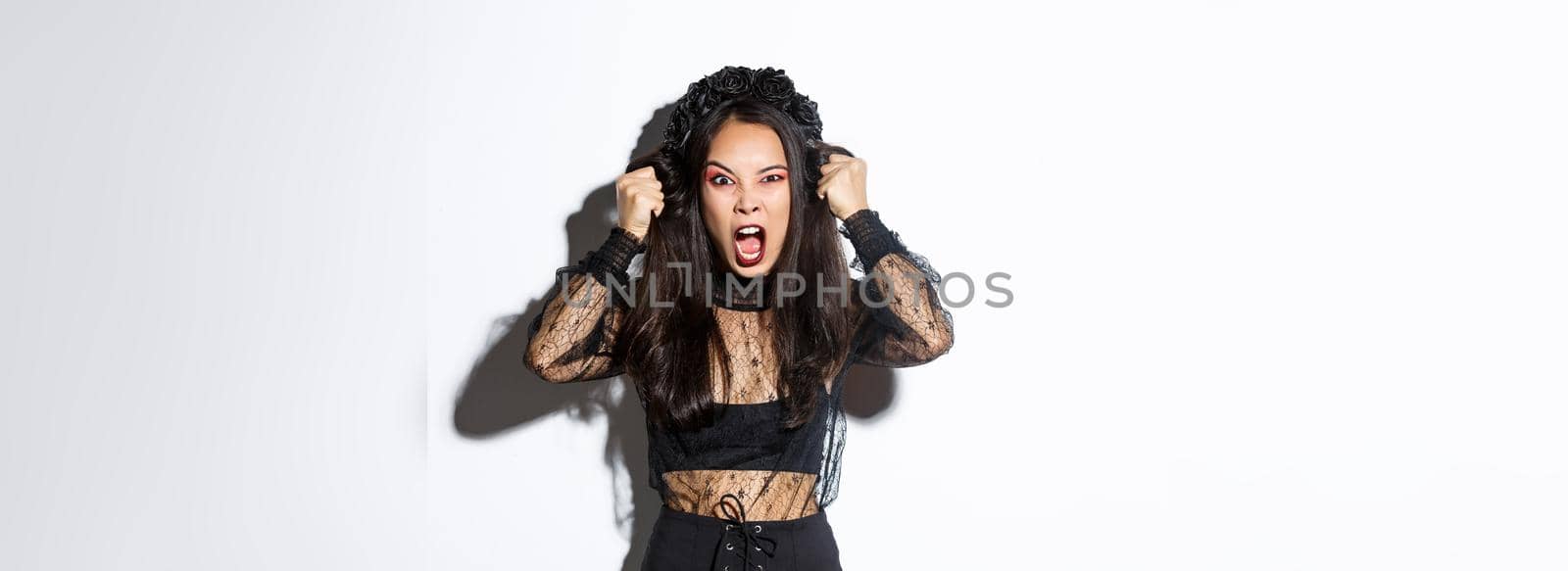 Angry asian woman impersonate evil witch on halloween party, looking mad and furious, screaming to scare people, standing over white background in gothic dress.