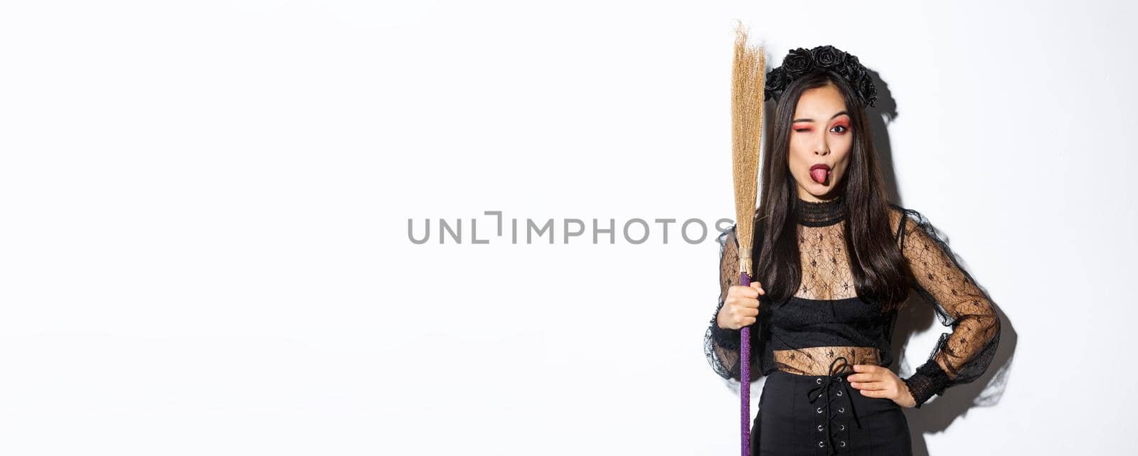 Beautiful asian woman celebrating halloween in witch costume, holding broom and showing tongue, winking silly at camera, standing over white background by Benzoix