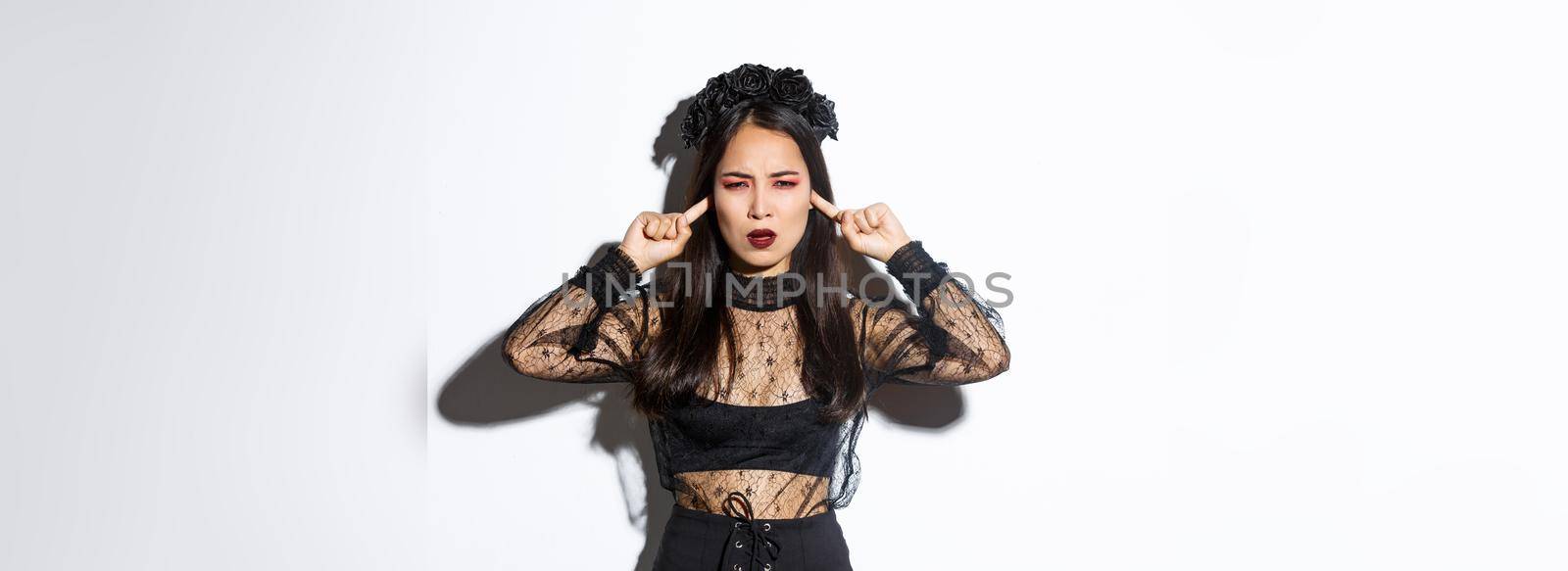 Annoyed and mad asian woman in witch halloween costume shut ears and frowning angry, standing bothered by something loud, complaining on noise over white background.