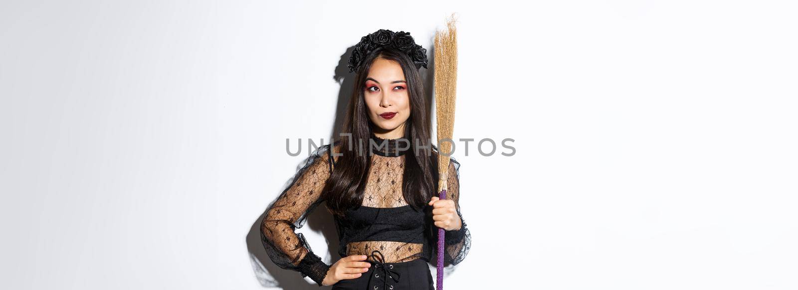 Portrait of smart thoughtful asian girl looking at upper left corner with pleased smirk, holding broom, wearing witch costume for halloween party, standing over white background by Benzoix