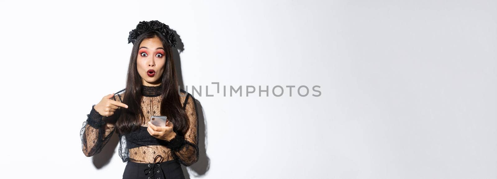 Portrait of amazed asian girl pointing finger at smartphone, showing something about halloween in internet, standing over white background in gothic lace dress.