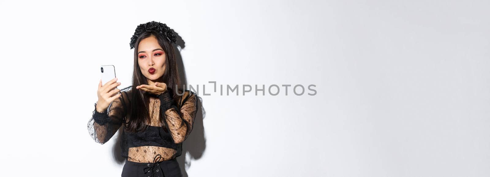 Portrait of stylish asian female blogger with gothic makeup and halloween costume sending air kiss at mobile phone camera, record video or having videocall, standing over white background.