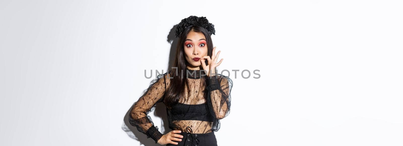 Pretty asian girl in witch costume making promise, seal lips and keep secret, zip mouth, censored information, standing over white background in halloween outfit by Benzoix