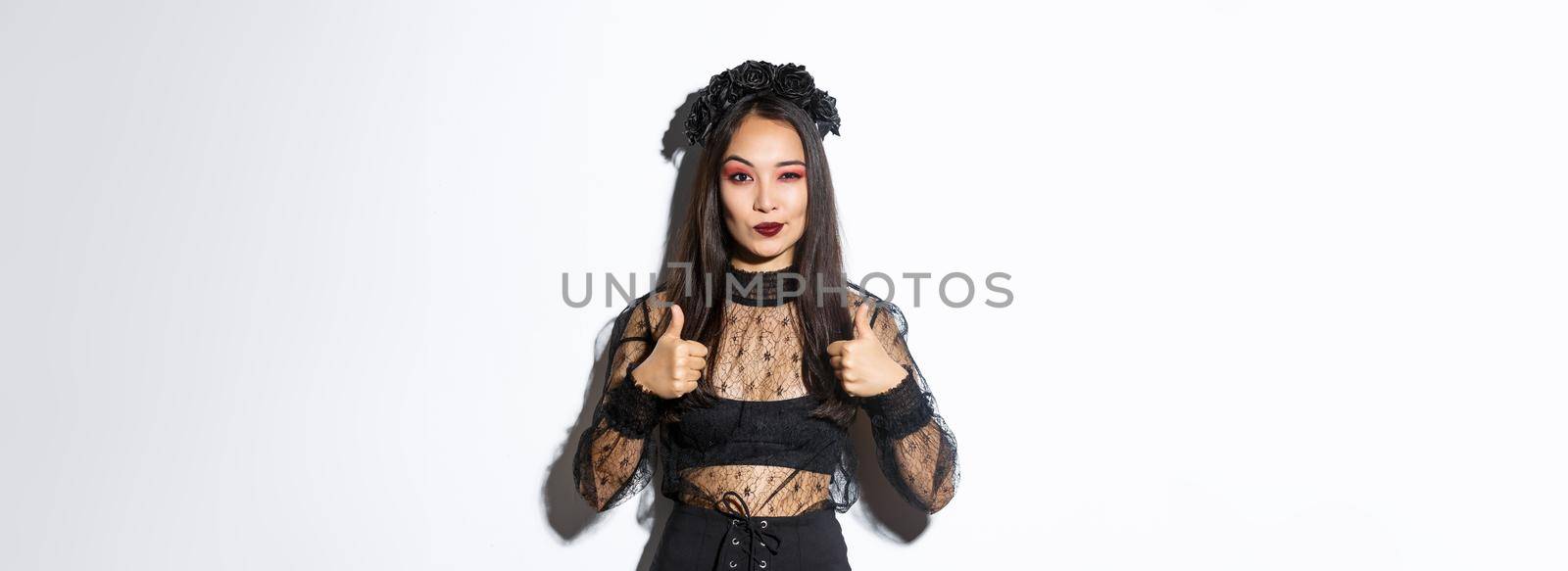 Pleased sassy attractive woman in witch dress showing thumbs-up in approval, like and praise your choice, making compliment, standing over white background by Benzoix