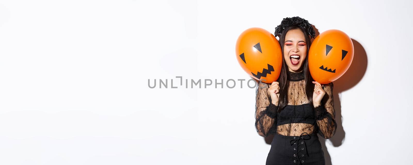 Image of asian girl in evil witch costume holding two orange balloons with scary faces, celebrating halloween, standing over white background by Benzoix