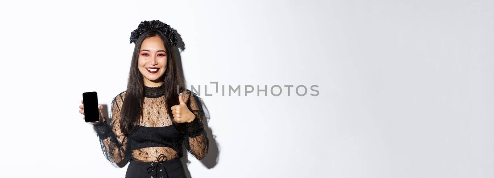 Image of happy and satisfied asian woman in halloween costume showing thumbs-up and demonstrating mobile phone screen, smiling pleased, standing over white background by Benzoix