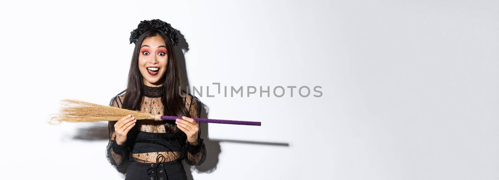 Excited young asian girl in witch costume looking amazed, showing small broom, standing over white background.