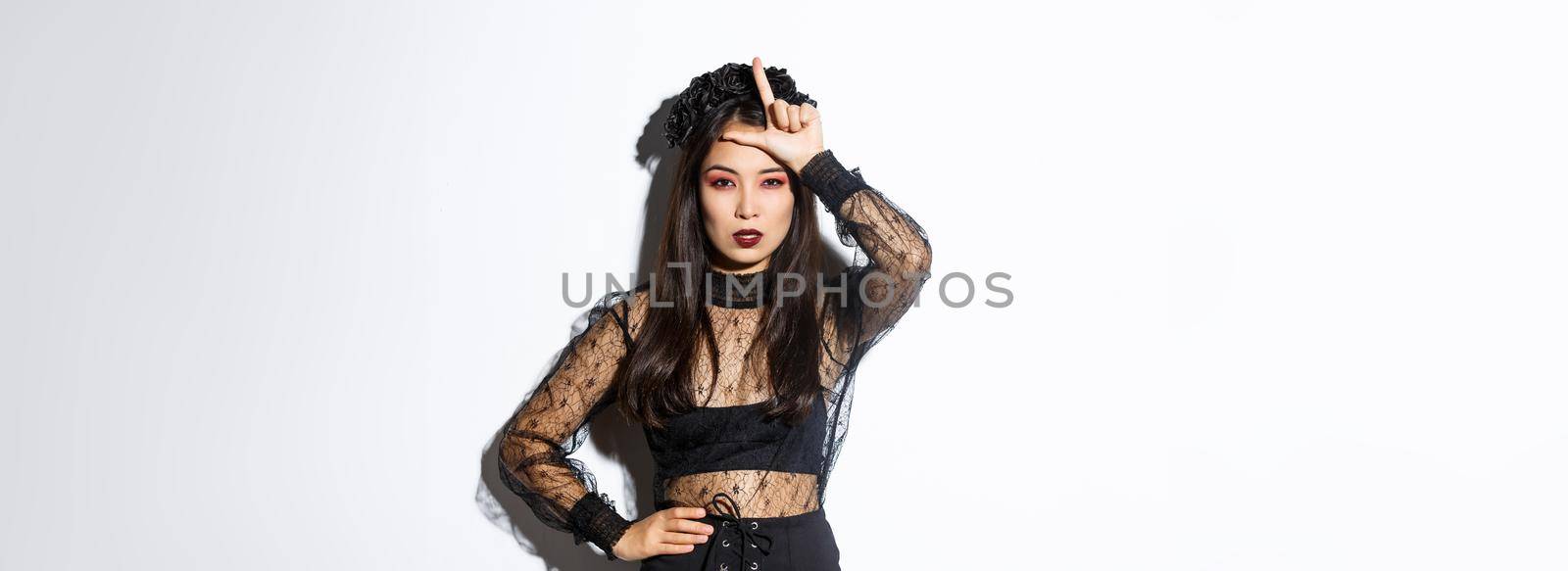 Image of arrogant sexy asian woman in witch costume showing loser gesture on forehead, mocking someone at halloween party, standing over white background by Benzoix