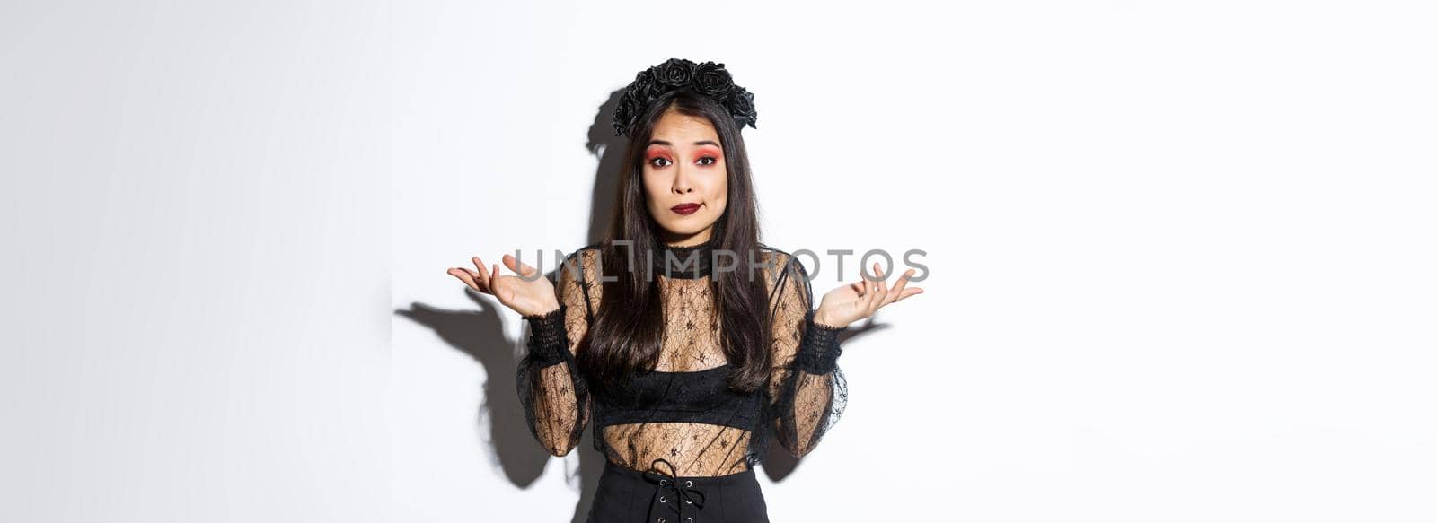 Image of clueless and unbothered asian girl looking confused, shrugging while standing over white background in halloween costume, dressed-up like evil witch on party by Benzoix