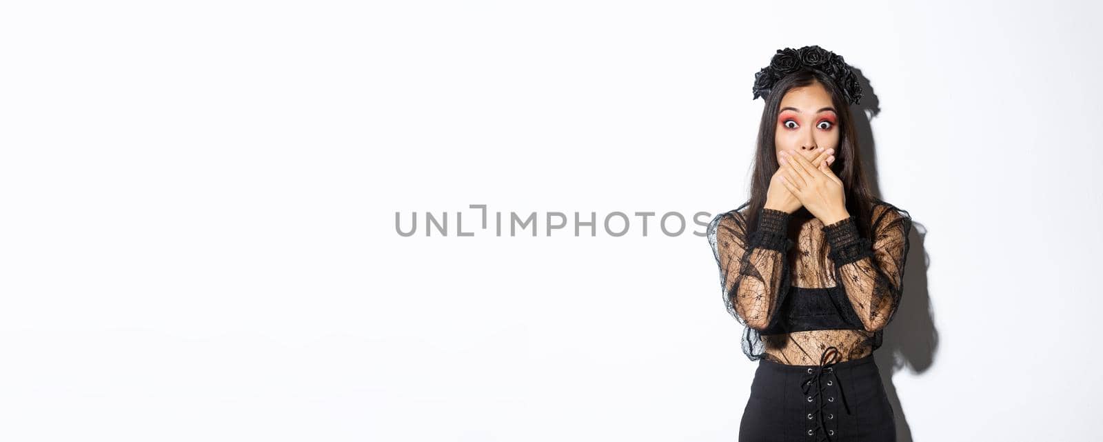 Shocked woman in witch outfit gasping and cover mouth speechless, standing over white background, looking at something surprising, wearing black lace dress and wreath, white background.