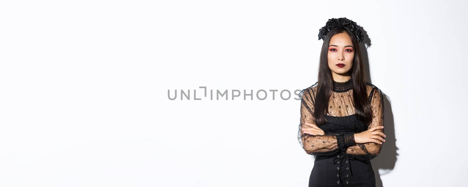 Image of beautiful asian woman in black lace dress and wreath looking serious. Girl dressed-up for halloween party as evil witch, standing over white background.
