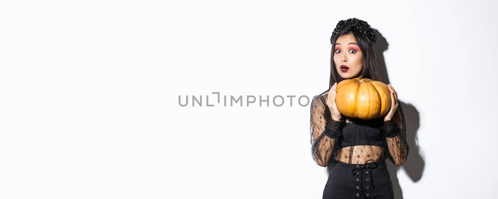 Portrait of woman lifting heavy pumpkin, getting ready for halloween, wearing witch costume, standing over white background.