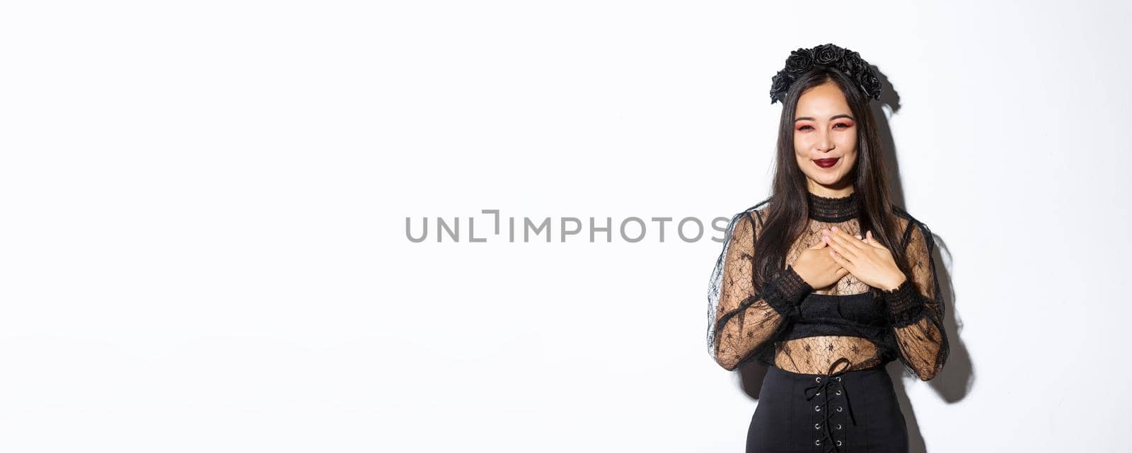 Pleased beautiful asian woman in witch costume smiling, holding hands on chest and looking grateful, express gratitude, standing in halloween outfit over white background.