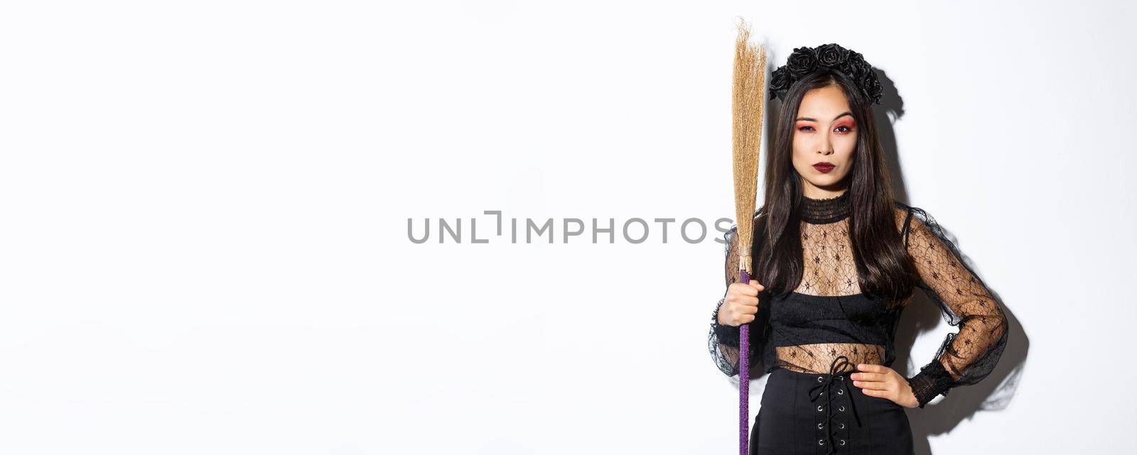 Beautiful asian woman in gothic lace dress and black wreath, holding broom and looking suspicious at camera, standing over white background by Benzoix