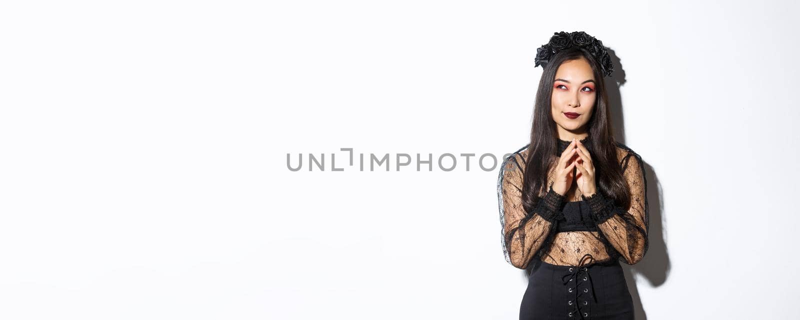 Thoughtful and cunning asian woman in witch costume prepared something for halloween, looking upper left corner and thinking. Girl wearing black gothic dress and wreath for party by Benzoix