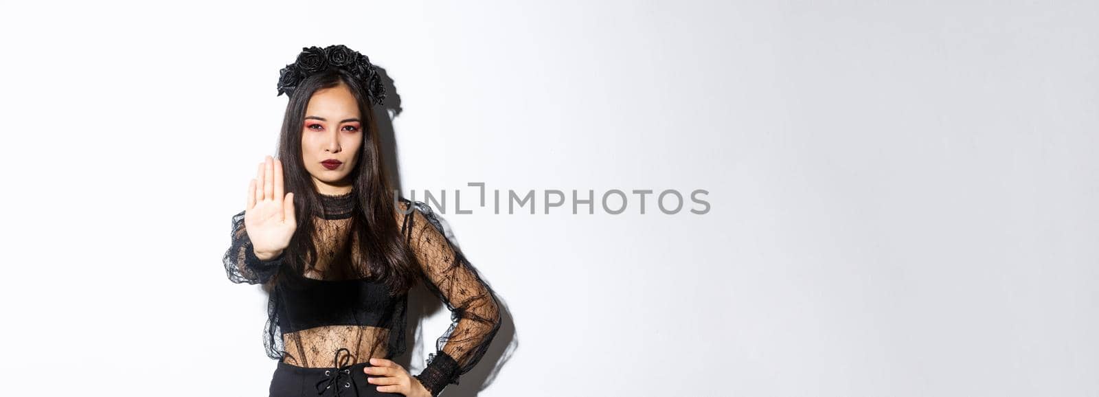 Image of serious asian woman in halloween costume of witch, showing stop gesture, forbid or prohibit something with displeased confident face, standing white background by Benzoix