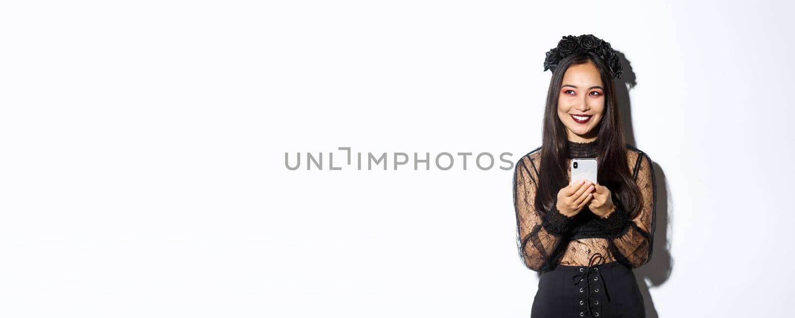Portrait of cunning beautiful asian woman, witch in gothic lace dress using mobile phone, smiling and looking at upper left corner, standing over white background.