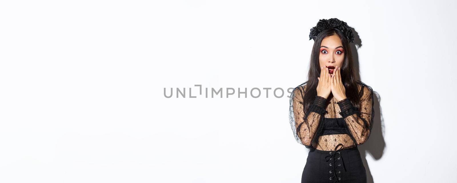 Surprised beautiful asian woman in witch costume looking amazed. Female wearing halloween gothic dress and wreath, open mouth aamused, standing over white background.