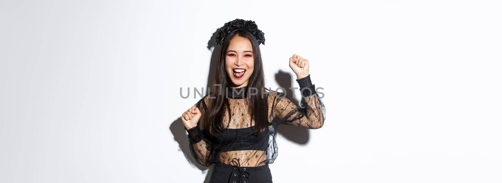 Image of young asian woman having fun on halloween party, raising hand up as rejoicing or triumphing, wearing witch gothic dress, standing over white background.