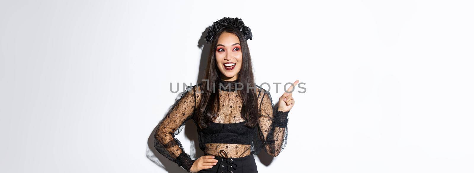 Excited happy asian woman in black lace dress and wreath looking amazed at upper left corner, pointing finger at your halloween promo banner, standing over white background by Benzoix
