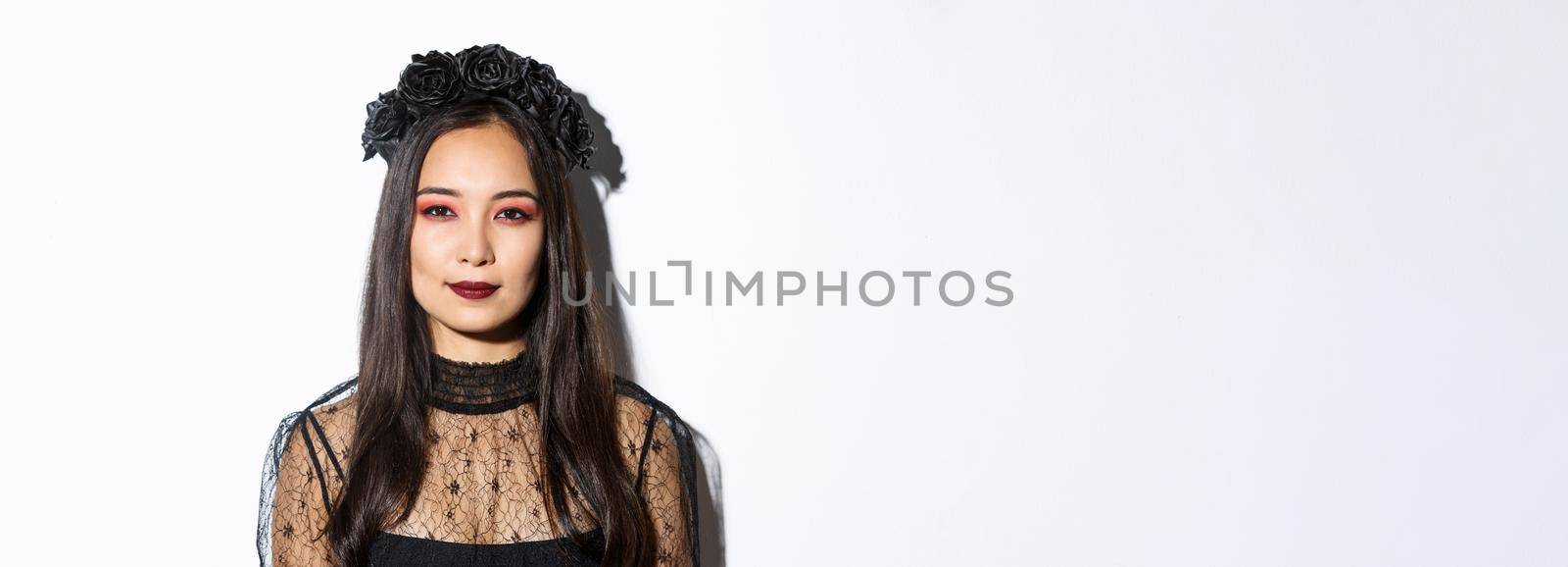 Close-up of beautiful gothic girl with black wreath, getting dressed for halloween party, standing over white background in wicked witch costume.