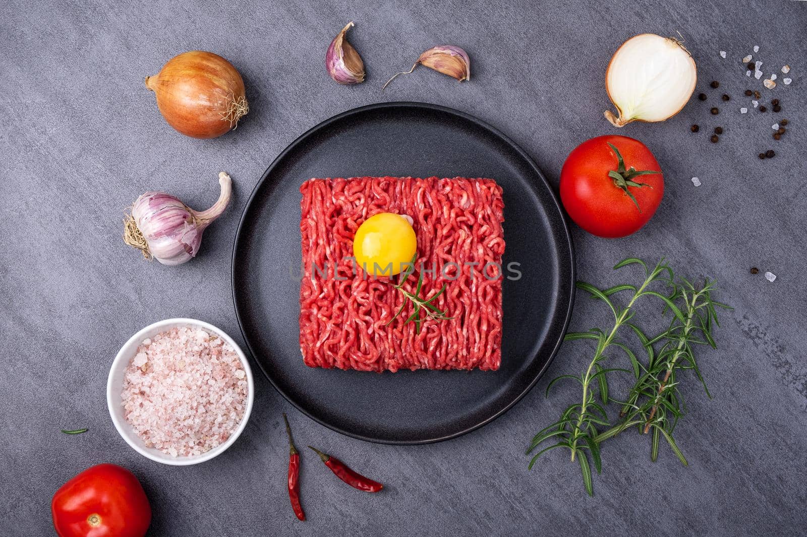 Minced meat lies on a black ceramic plate surrounded by cooking ingredients. View from above. Selective focus.