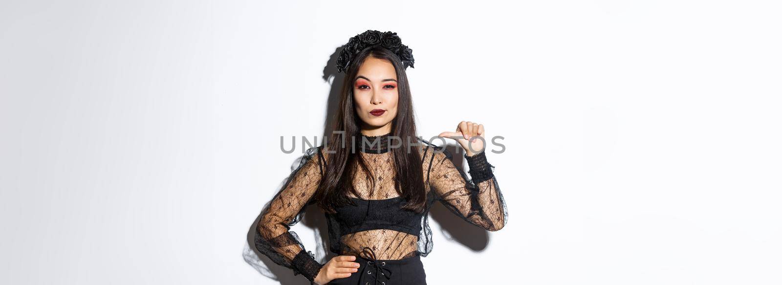 Confident stylish asian woman looking determined, wearing black lace dress for halloween party, pointing at herself sassy, standing over white background.