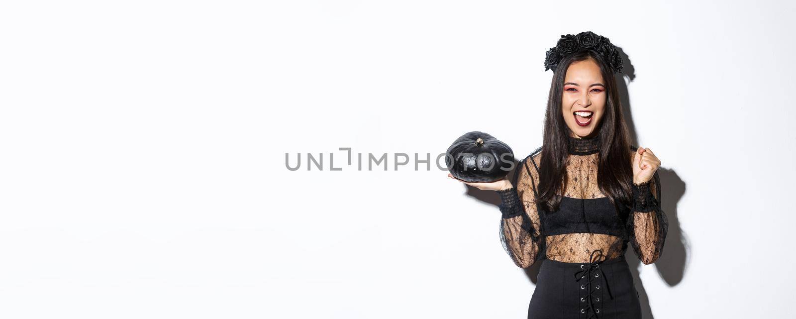 Attractive joyful asian girl enjoying halloween, holding black pumpkin and cheering, wearing witch costume, standing over white background by Benzoix