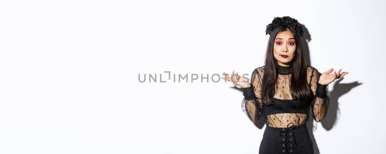 Image of clueless and unbothered asian girl looking confused, shrugging while standing over white background in halloween costume, dressed-up like evil witch on party.