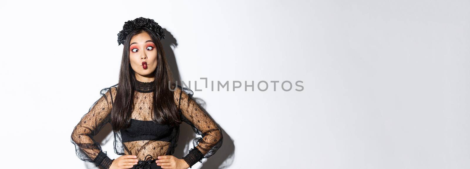 Image of funny asian girl celebrating halloween, wearing gothic lace dress to impersonate witch, showing silly faces and having fun, standing over white background.