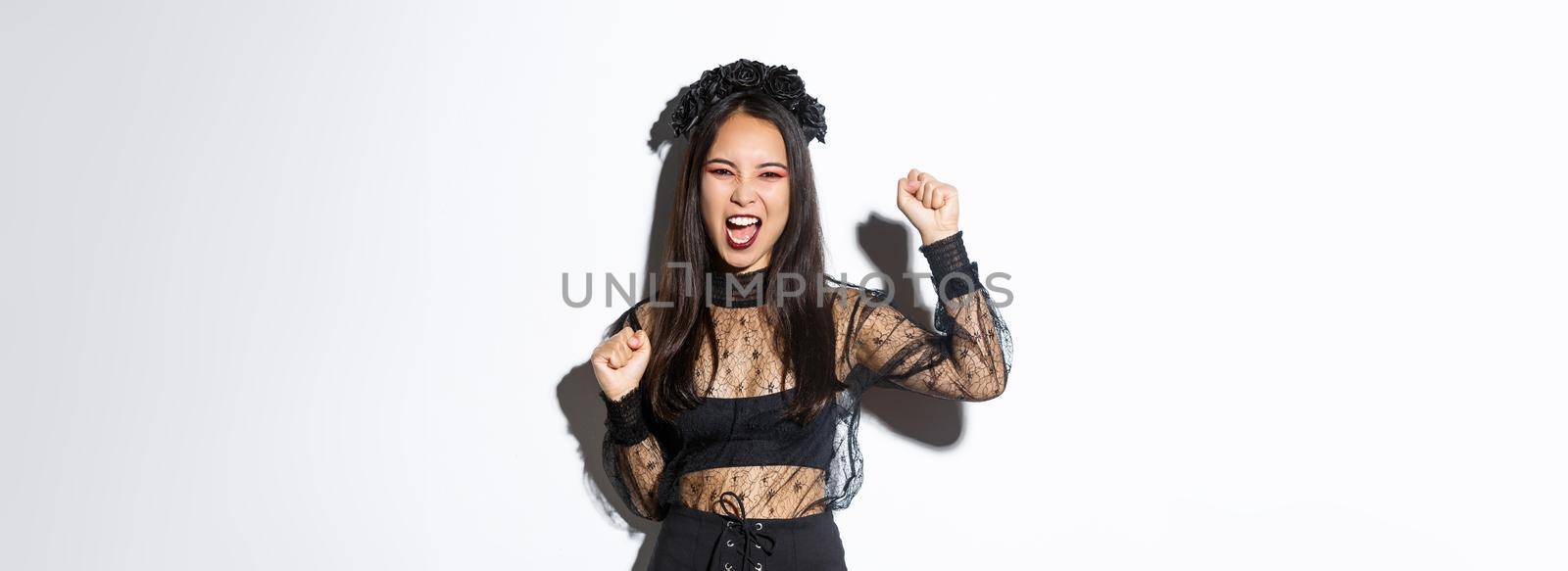 Excited asian woman in elegant gothic lace dress and wreath celebrating halloween, enjoying party, raising hand up and yelling joyful, standing over white background.