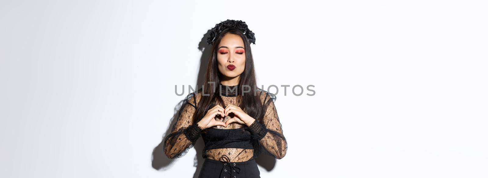 Image of beautiful asian gothic girl close eyes and pouting for kiss, showing heart gesture, express love or sympathy on halloween party, standing over white background.