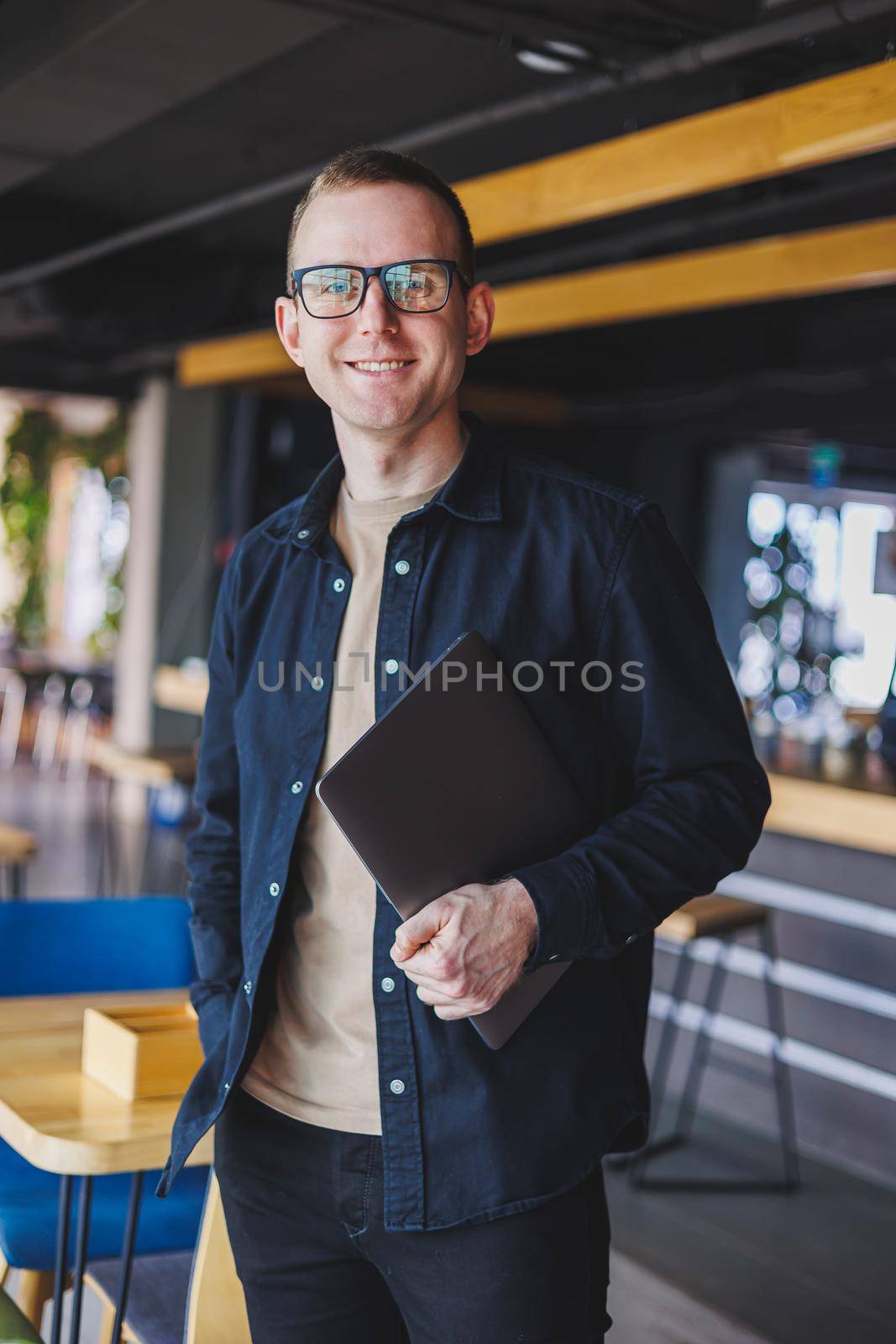 Male entrepreneur in black shirt and glasses standing in office with laptop in hand, successful corporate boss feeling good from rich lifestyle
