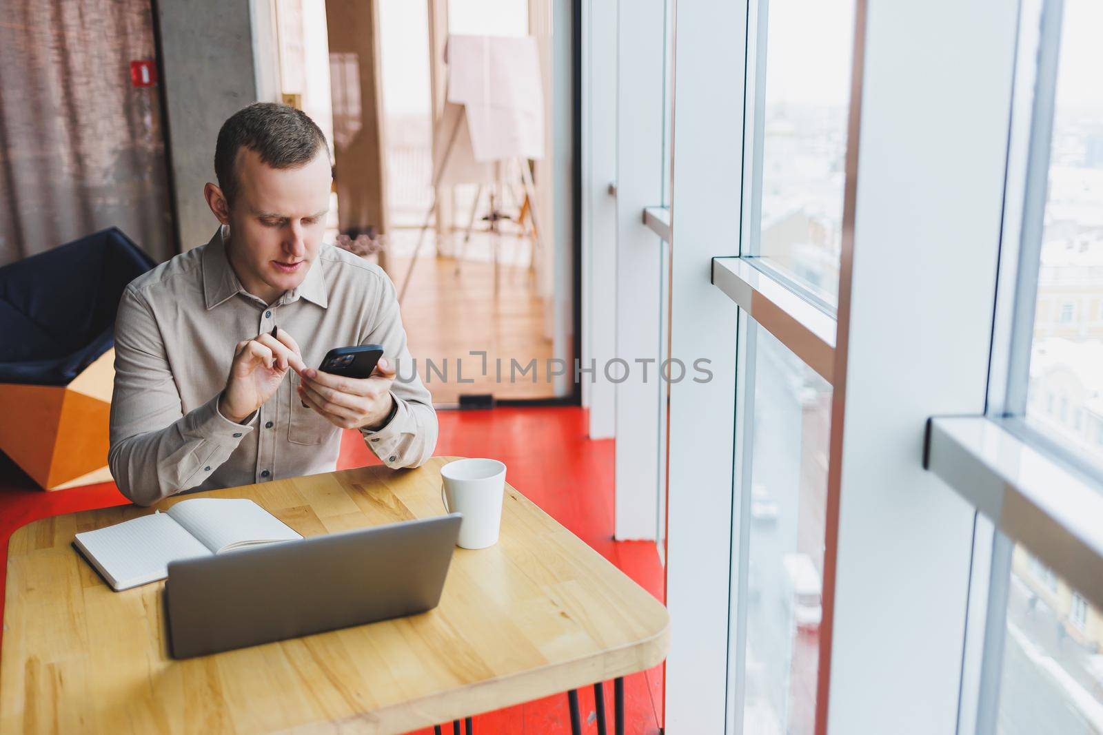 Successful young man is sitting at a table in a cafe-restaurant indoors, working and studying on a laptop computer,holding a mobile phone with a blank screen. Freelancer mobile office business concept