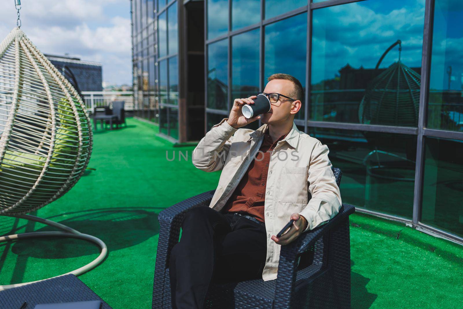 Confident young businessman, dressed in formal wear and stylish glasses, calls on a smartphone while sitting with coffee, on the terrace of a modern office building with a glass wall