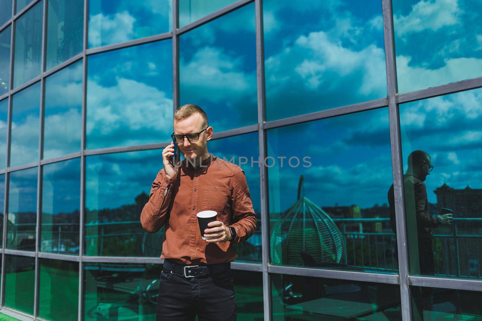 Attractive fashionable male businessman in a stylish shirt and trousers talking on the phone near the office building and drinking coffee, modern glass office