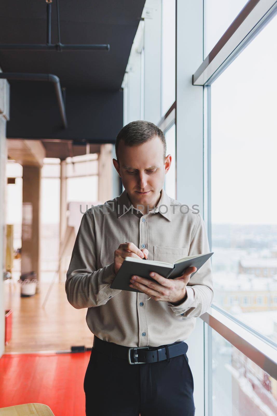A young businessman stands at a panoramic window in a skyscraper and makes notes in his notebook with a pen. A man in a shirt and trousers of European appearance