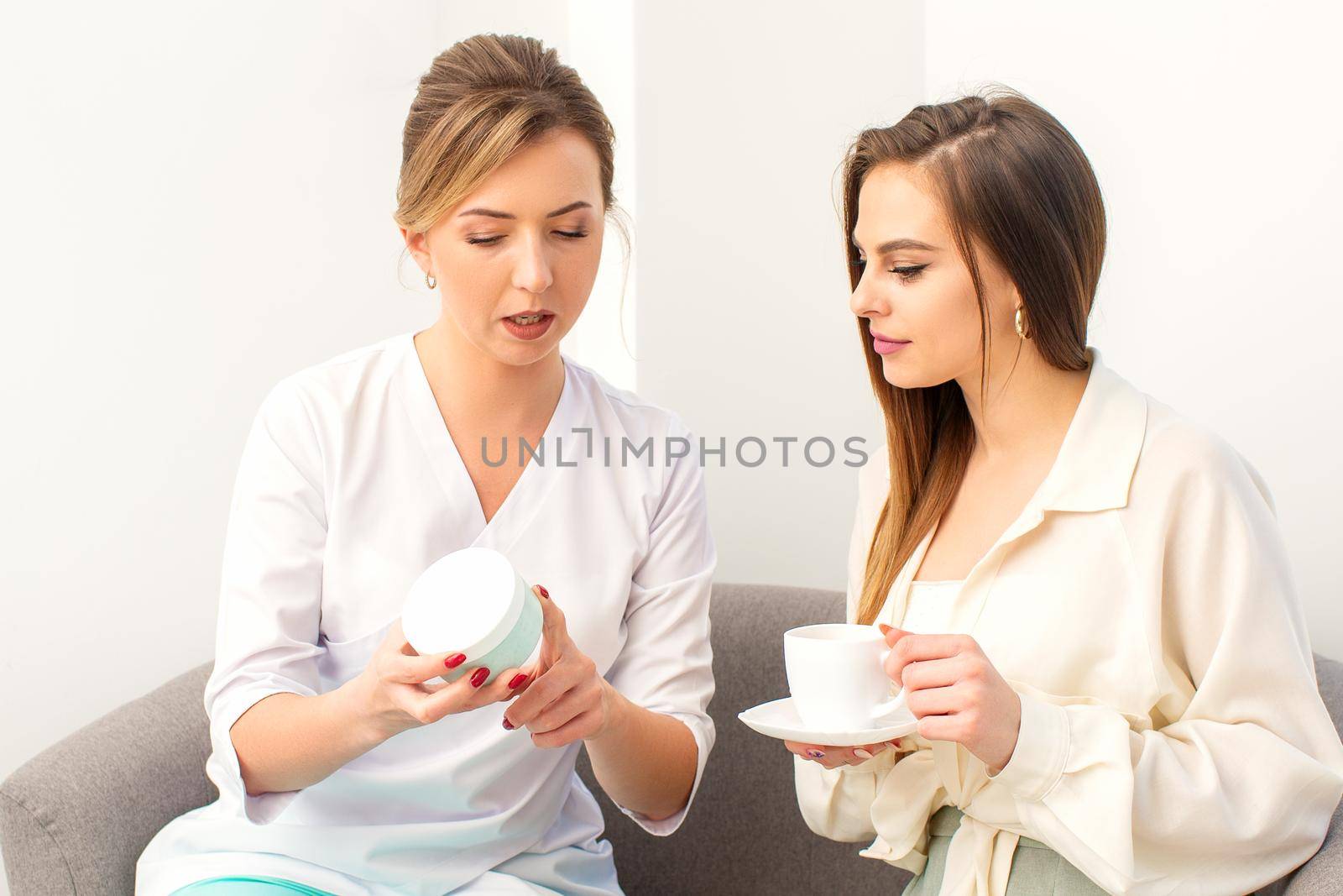 Beautician offering product for the young woman holding a white plastic jar with a cream sitting on the sofa