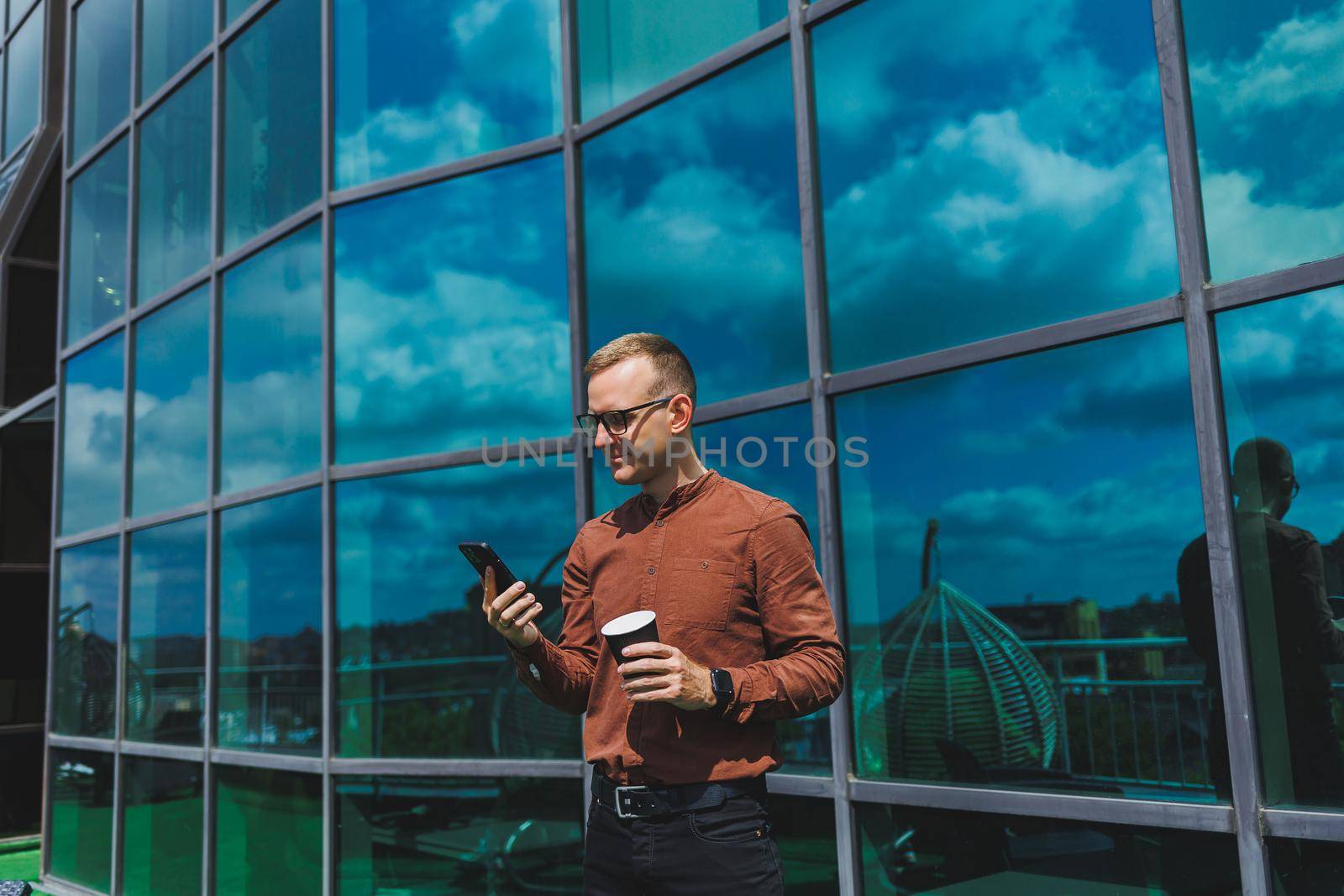 Attractive fashionable male businessman in a stylish shirt and trousers talking on the phone near the office building and drinking coffee, modern glass office