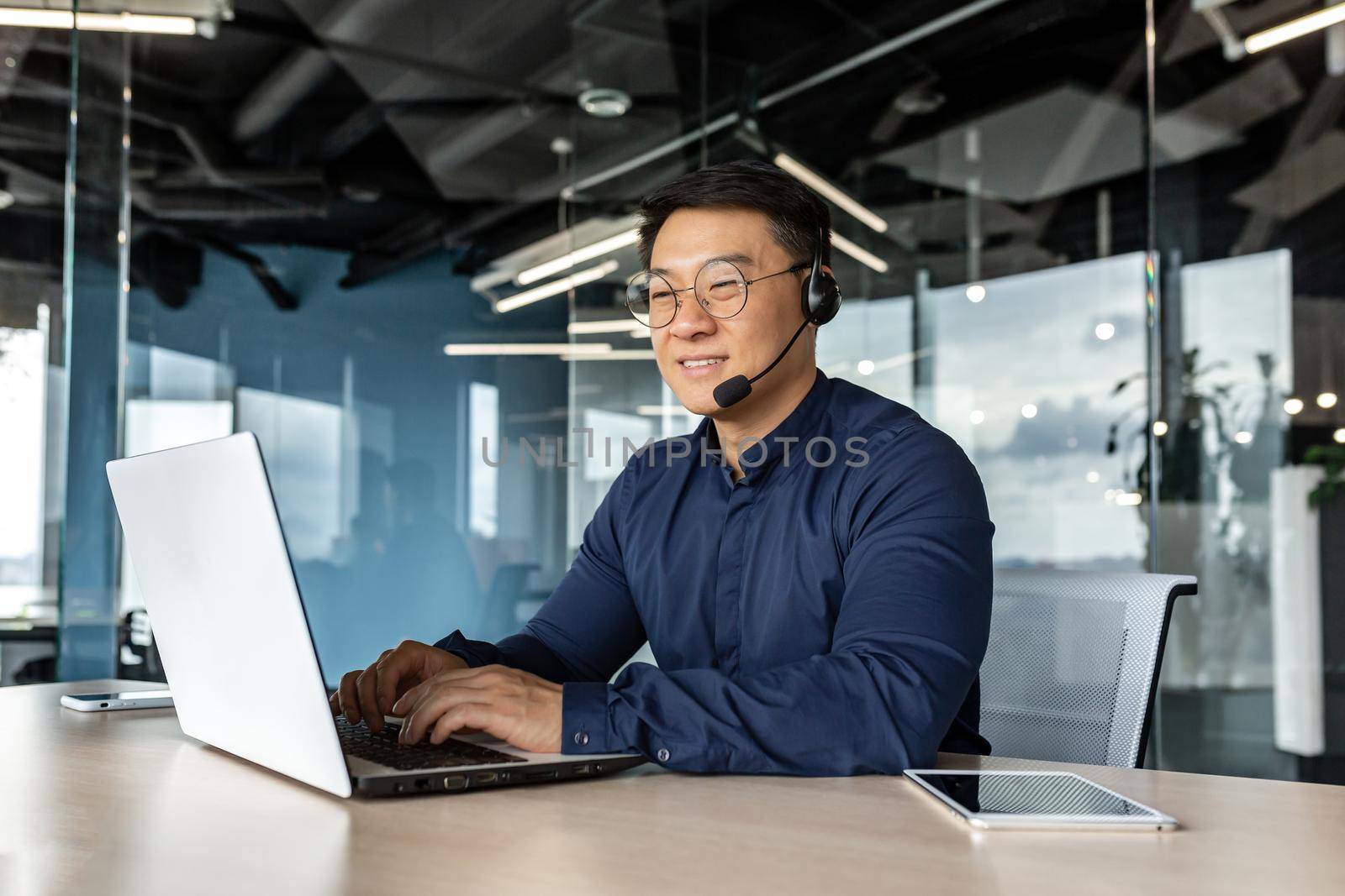 Happy and smiling worker with headset for video call, Asian tech support man talking to online service users, businessman using laptop for online customer support.