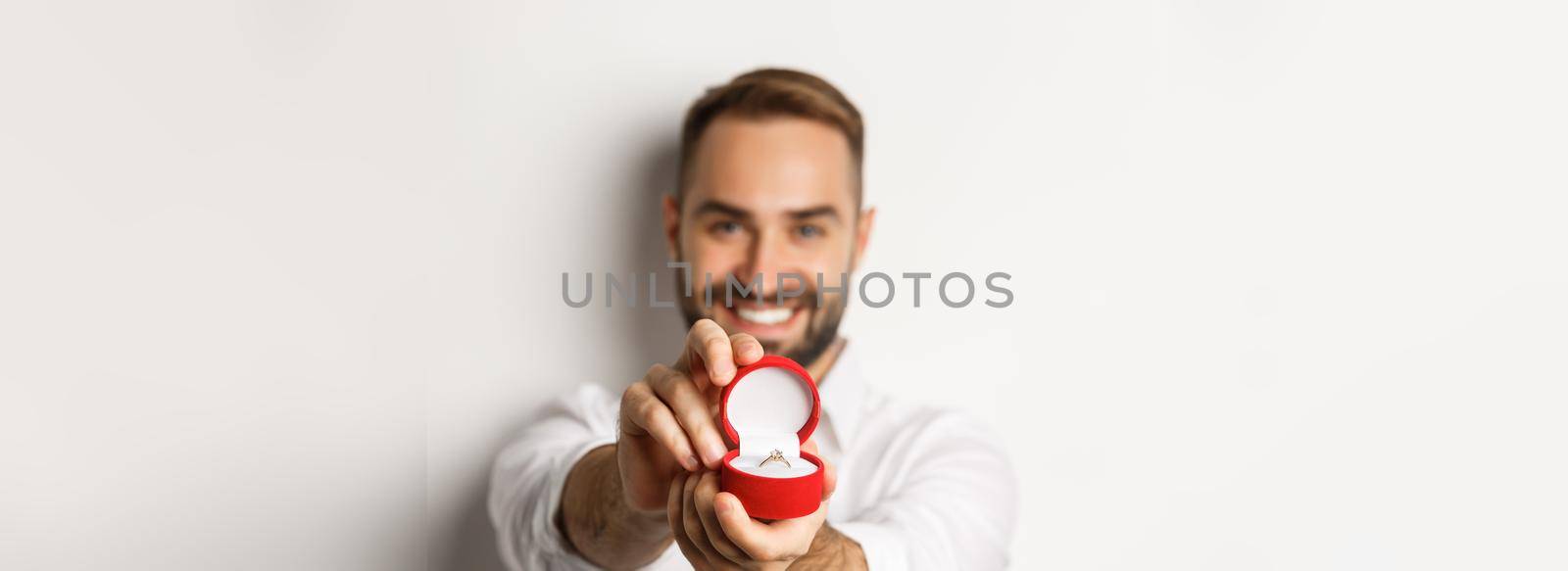 Close-up of handsome man asking to marry him, focus on box with wedding ring, concept of proposal and relationship, white background.